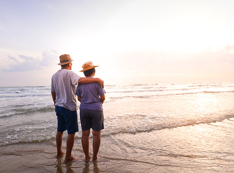 A bare-footed couple stands arm in arm in the wet sand by the ocean, watching the sunset.