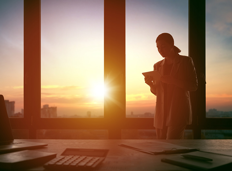 A woman standing by an office window, the sun setting outside